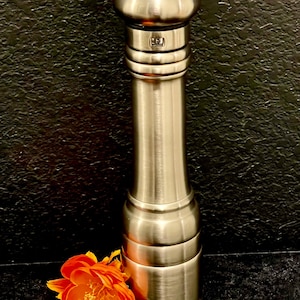 May include: A stainless steel pepper mill with a rounded top and a cylindrical body. The mill has a brushed metal finish and is accompanied by an orange and yellow flower. The pepper mill is a kitchen utensil.