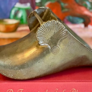 May include: A vintage brass shoe horn with a seashell design. The shoe horn is a warm, metallic color and sits atop a red book. The seashell is embossed on the top of the shoe horn.