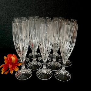 May include: A collection of clear, ribbed champagne flutes with a decorative base. The glasses are arranged on a black surface, with an orange and yellow flower in the foreground. The flutes are made of glass.