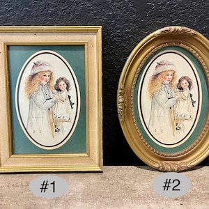 Könnte beinhalten: Zwei gerahmte Vintage-Drucke. Einer ist rechteckig, der andere oval. Jeder zeigt eine Aquarellillustration eines Mädchens, das eine Puppe hält, vor einem grünen Hintergrund. Die Rahmen sind goldfarben und verleihen einen klassischen Touch.