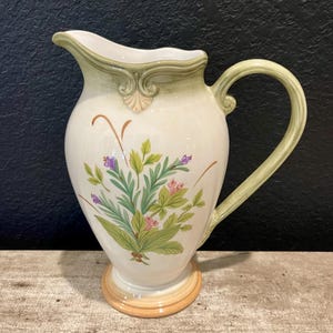 May include: A white ceramic pitcher with a green handle and rim. The pitcher features a floral design with green leaves, purple and pink flowers, and brown stems. The base is a light orange color.