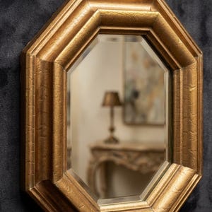 May include: An octagonal wall mirror featuring a textured, metallic gold frame with a bevelled glass edge. The frame has a prominent, multi-faceted border and includes a small, centred gold hanging loop at the top for wall mounting.