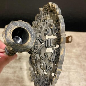 May include: Ornate, antique-style candle holder in a dark bronze finish. The holder features intricate floral and scrollwork designs. The candle cup is circular, and the piece has a small loop for hanging. The overall aesthetic is vintage and decorative.