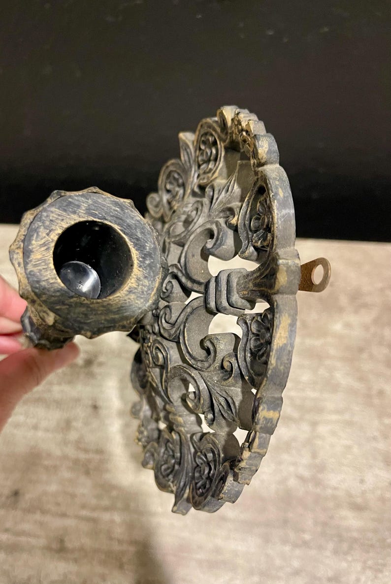 May include: Ornate, antique-style candle holder in a dark bronze finish. The holder features intricate floral and scrollwork designs. The candle cup is circular, and the piece has a small loop for hanging. The overall aesthetic is vintage and decorative.