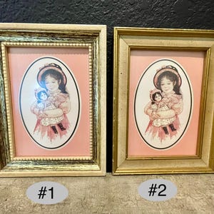 Könnte beinhalten: Zwei gerahmte Vintage-Drucke. Jeder zeigt ein ovales Porträt eines Mädchens in einem rosa Kleid, das eine Puppe hält. Die Rahmen sind rechteckig, einer goldfarben und der andere cremefarben und goldfarben. Der Hintergrund ist schwarz.
