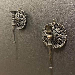 May include: Two ornate, antique-style wall sconces. Each sconce features a candle holder with a long, tapered stem and a decorative, circular backplate with swirling floral patterns. The sconces are made of a dark metal with a distressed finish, mounted on a dark gray wall.