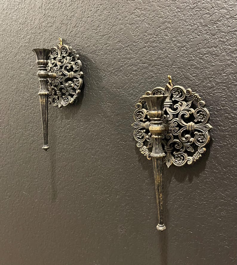 May include: Two ornate, antique-style wall sconces. Each sconce features a candle holder with a long, tapered stem and a decorative, circular backplate with swirling floral patterns. The sconces are made of a dark metal with a distressed finish, mounted on a dark gray wall.