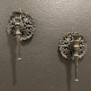 May include: Two ornate wall sconces with a dark metallic finish. Each sconce features a central candle holder and a decorative circular backing with floral and scrollwork details. The sconces are mounted on a textured, dark gray wall.