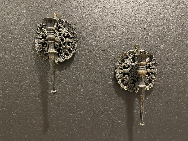 May include: Two ornate wall sconces with a dark metallic finish. Each sconce features a central candle holder and a decorative circular backing with floral and scrollwork details. The sconces are mounted on a textured, dark gray wall.
