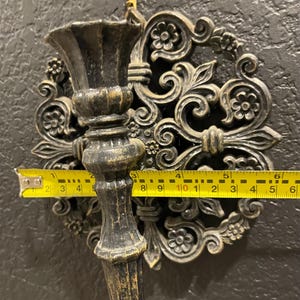 May include: Ornate, dark bronze wall sconce with a floral design. The sconce measures approximately 6 inches wide. The sconce has a decorative backplate and a cup-shaped holder for a candle or other item. The sconce is mounted on a dark textured wall.