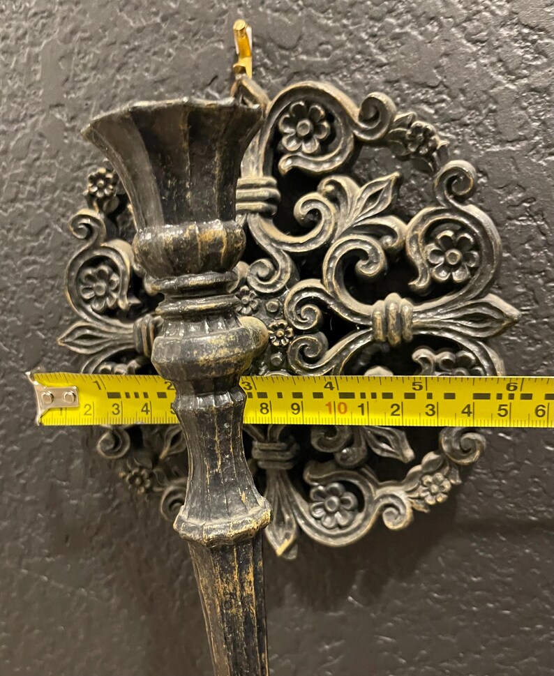 May include: Ornate, dark bronze wall sconce with a floral design. The sconce measures approximately 6 inches wide. The sconce has a decorative backplate and a cup-shaped holder for a candle or other item. The sconce is mounted on a dark textured wall.