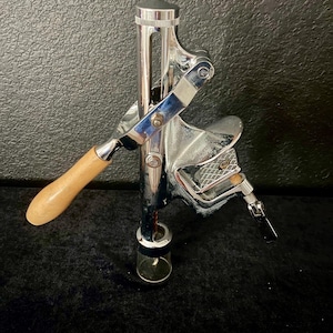 May include: A chrome-plated metal wine bottle opener featuring a vertical lever mechanism, a light-coloured wooden handle, and a clamp-style base with a textured grip. The device includes a central shaft, a metal worm, and a small circular base ring.