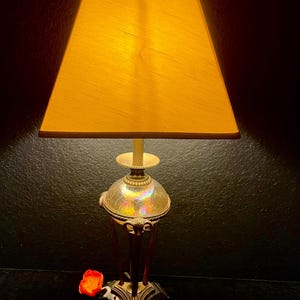 May include: A vintage table lamp with a square, yellow lampshade. The lamp base has a decorative, iridescent glass globe and a dark, ornate base. A small, red flower sits near the base of the lamp.