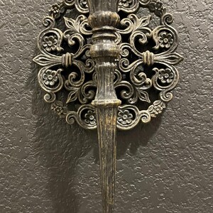 May include: Ornate, antique-style wall sconce with a dark bronze finish. The sconce features a central torch-like holder and is surrounded by a decorative, floral-patterned circular frame. The design evokes a vintage aesthetic, suitable for interior decor.