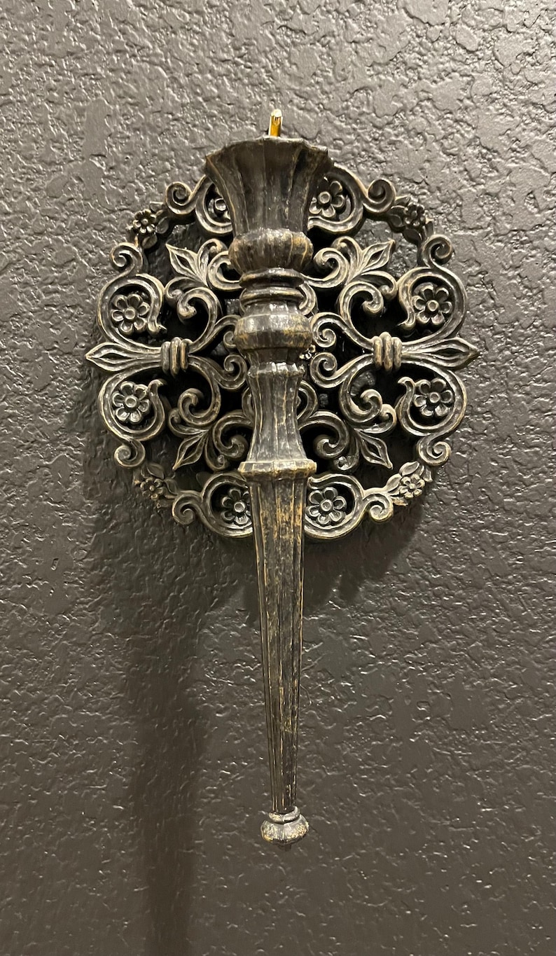 May include: Ornate, antique-style wall sconce with a dark bronze finish. The sconce features a central torch-like holder and is surrounded by a decorative, floral-patterned circular frame. The design evokes a vintage aesthetic, suitable for interior decor.