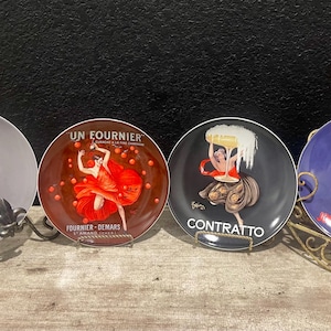 Può includere: Quattro piatti decorativi con illustrazioni in stile vintage. I piatti presentano immagini di donne, champagne e testo. I piatti sono esposti su supporti metallici decorativi, con un supporto dorato a forma di gallo a destra.