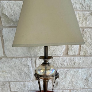 May include: A vintage table lamp with a beige lampshade and a decorative brass base. The lamp sits on a round, dark wood table with ornate detailing. The lamp's glass globe has an iridescent finish.