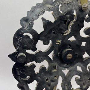 May include: Close-up of a dark, ornate decorative piece with floral and scrollwork designs. The piece has a circular shape with the text "HOMCO MADE IN USA 4149" and "COPYRIGHT MCMLXXIII" visible. The material appears to be a dark, possibly metallic, composite.