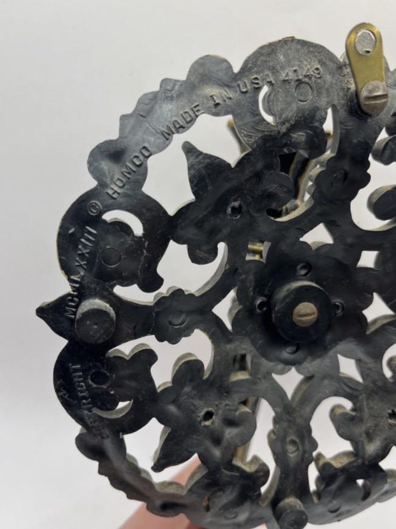 May include: Close-up of a dark, ornate decorative piece with floral and scrollwork designs. The piece has a circular shape with the text "HOMCO MADE IN USA 4149" and "COPYRIGHT MCMLXXIII" visible. The material appears to be a dark, possibly metallic, composite.