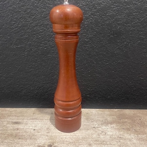 Puede incluir: Un molinillo de pimienta de madera, de color marrón rojizo. Tiene una parte superior redondeada con un botón plateado y un cuerpo cilíndrico. La base está detallada con varios anillos decorativos. Mide unos 20 cm de altura.