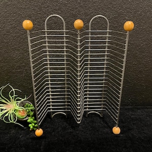 May include: A silver-toned metal wire rack featuring a zigzag design with multiple horizontal slots for storage. The structure is topped and bottomed with small, round, light-colored wooden spheres at the ends of the vertical support wires.