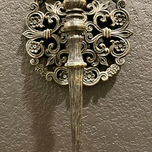 May include: Ornate, antique-style wall sconce with a dark bronze finish. The sconce features a central torch-like holder and a decorative circular backing with floral and scrollwork details. A classic piece for home decor.