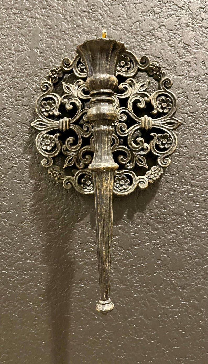 May include: Ornate, antique-style wall sconce with a dark bronze finish. The sconce features a central torch-like holder and a decorative circular backing with floral and scrollwork details. A classic piece for home decor.
