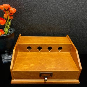 May include: A wooden desk organizer with a warm brown finish, featuring a single drawer with a metal label pull and an upper tier with four circular cutouts for cable management. The organizer has a tiered design with a flat top surface.