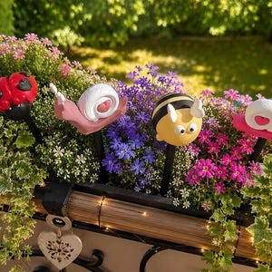 Könnte beinhalten: Vier Gartenstecker mit bunten Insekten- und Weichtierfiguren: ein roter Marienkäfer, zwei rosa Schnecken mit weißen Häusern und eine gelbe Biene mit schwarzen Streifen. Jede Figur ist auf einem dünnen schwarzen Stab zur Befestigung montiert.