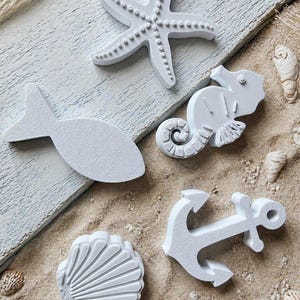 Könnte beinhalten: Weiße Dekorationsobjekte zum Thema Nautik, darunter ein Seestern, ein Fisch, ein Seepferdchen, ein Anker und eine Muschel. Auf Sand mit Muscheln und einem verwitterten Holzbrett angeordnet.