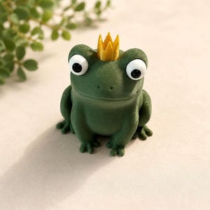 Könnte beinhalten: Eine kleine, dunkelgrüne Froschfigur mit strukturierter Oberfläche, großen, runden, weißen Augen mit schwarzen Pupillen und einer kleinen, spitzen gelben Krone auf dem Kopf. Der Frosch befindet sich in einer sitzenden Position.