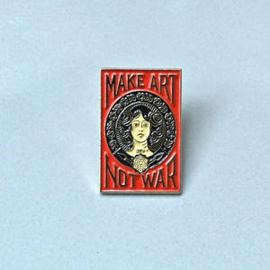 Op de afbeelding: Rode emaille pin met een zwart-witte illustratie van een vrouw met een bloemenkrans en de tekst "Make Art Not War".