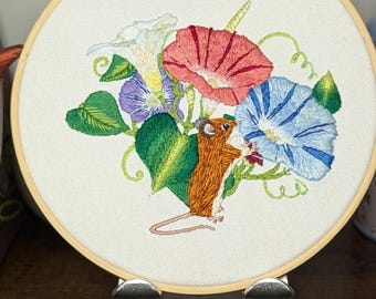 May include: Embroidered hoop art featuring a brown mouse with a red hat, surrounded by colorful morning glory flowers in shades of red, blue, purple, and white. The artwork is set against a cream-colored fabric background, framed by a wooden hoop.