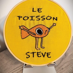 May include: A yellow embroidery hoop with a cartoon fish with the text "LE POISSON STEVE" embroidered on it.