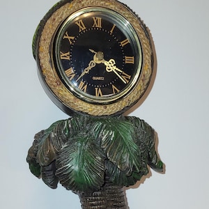 May include: A decorative clock featuring a black clock face with Roman numerals, set atop a palm tree design. The base showcases three monkey figurines, each in a 'see no evil' pose. The clock is approximately 10 inches tall.