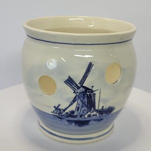 May include: A cream-colored ceramic candle holder with a rounded body and a flared rim. The exterior features a blue painted illustration of a traditional windmill near water, accented by circular cutouts and a blue band around the base.