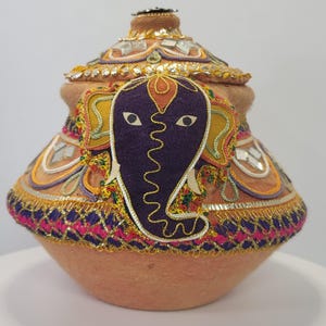 May include: A decorative, handcrafted pot with a lid, featuring an elephant design. The pot is primarily peach-colored, adorned with intricate embroidery in gold, purple, yellow, and pink. The lid is embellished with mirrors and a decorative top.