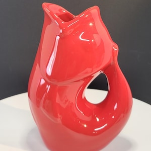 May include: A bright red ceramic pitcher shaped like a stylized fish. The vessel features a glossy finish, a wide open mouth at the top serving as a spout, and a curved handle that forms a circular opening on the side of the body, creating a smooth silhouette.