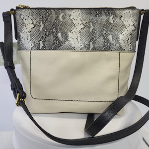 May include: A Fossil crossbody bag featuring a two-tone design with a grey and white snakeskin-patterned upper section and a solid cream-colored lower section. The bag includes a black adjustable shoulder strap, gold-tone hardware, and a zipper.