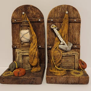 May include: Pair of nautical-themed bookends. Each bookend features a wooden door design with a lantern and fishing net on one, and an anchor and net on the other. The bookends are brown, with tan and orange accents.