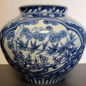 May include: A round, blue and white ceramic vase with a wide body and a narrow neck. The vase features a detailed floral and bamboo design in blue against a white background. The rim has a wave pattern.