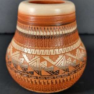 May include: A small, handcrafted ceramic vase with a rounded body and a cylindrical neck. The vase features intricate geometric patterns in shades of brown, cream, and black. The design includes triangles, lines, and other shapes, creating a textured appearance.