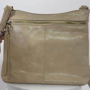 May include: A tan crossbody bag with a metallic sheen. The bag features a zippered main compartment and an exterior zippered pocket. It has a long, adjustable strap and a small tassel on the zipper pull. The bag is made of a soft, textured material.