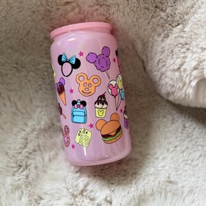 Pode incluir: Um copo rosa brilhante com uma tampa rosa a condizer. O copo é decorado com ícones de desenhos animados coloridos, incluindo orelhas do Mickey Mouse, um laço, gelado e um hambúrguer. O copo foi concebido para conter bebidas.