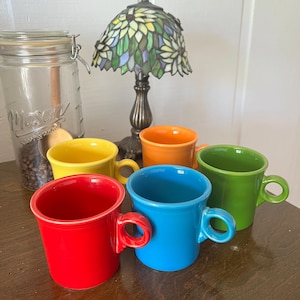 May include: A set of six brightly coloured ceramic mugs in red, yellow, orange, blue, and green. The mugs are displayed on a wooden surface, with a glass jar and a stained glass lamp in the background. The mugs have a classic design.