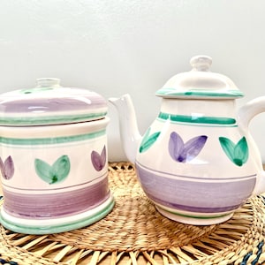 May include: A white ceramic teapot and sugar bowl set. The teapot has a curved spout and handle, with a lid. Both pieces are decorated with green and purple leaf designs and stripes. The set is placed on a woven mat.