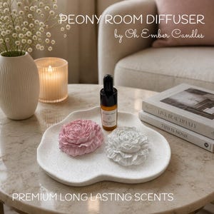 May include: A white speckled tray holds a pink peony-shaped diffuser, a white peony-shaped diffuser, and a small amber glass bottle with a black dropper. Text reads PEONY ROOM DIFFUSER by Oh Ember Candles and PREMIUM LONG LASTING SCENTS.