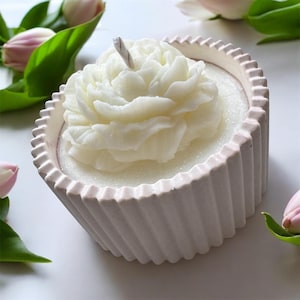 May include: A white, flower-shaped candle in a light pink, fluted container. The candle has a wick and is surrounded by white wax. Pink tulips and green leaves are in the background.