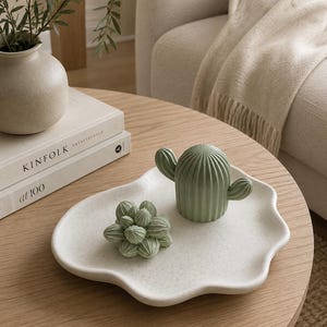 May include: A white speckled tray with an irregular, wavy edge holds two sage green decorative objects. One object is a ribbed, barrel-shaped cactus with two small arms, and the other is a cluster of small, rounded, textured succulent-like shapes.