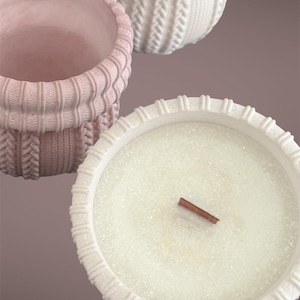 May include: Close-up of three decorative candle holders. One is a light pink, the other two are white. The candle holders have a textured, knitted sweater design. One candle has a wooden wick.
